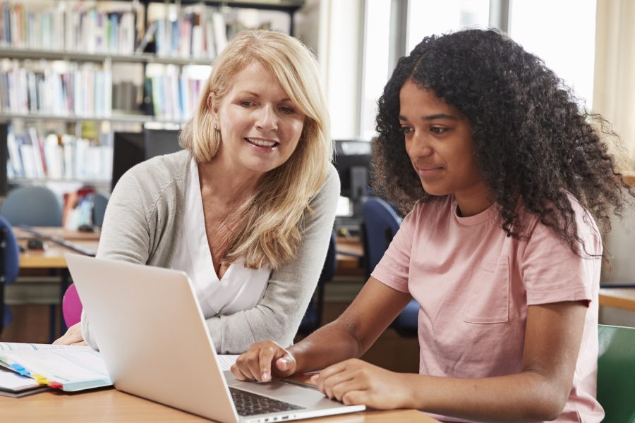 Een docent buigt zich over een laptop en wijst iets aan voor een leerling die meekijkt.